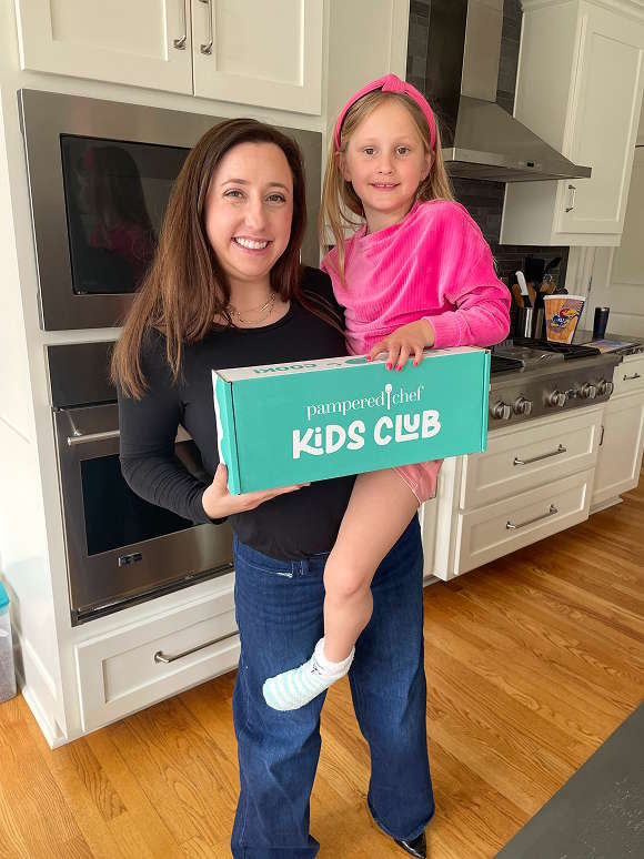 A young girl wearing the Kids Club Apron who’s dancing and holding a Kids Club box.